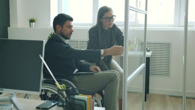 Disabled man working with sticky notes on glassboard and discussing project with female colleague in modern office. People and cooperation concept.