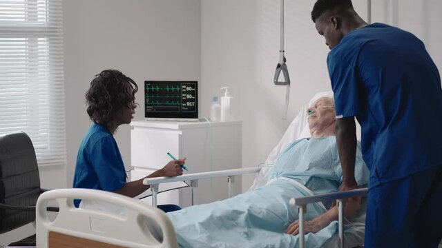 A team of african ethnic group doctors a man and a woman talk to an elderly male patient. A doctor and a nurse talk to a bedridden patient in a hospital ward