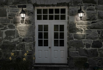 White metal doors and transom window on a grey natural stone building, illuminated by two lantern style exterior lights, nobody