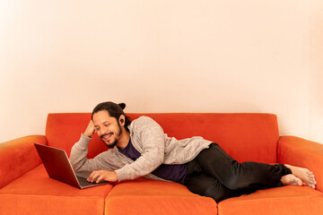 Fototapeta premium Young man dating online using the computer laptop laying on an orange couch sofa at home in the living room.