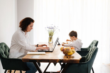 Middle age woman working at home on laptop while child is eating food on the same table with tablet. Working mother with son using gadgets on wooden table kitchen. Busy freelancer, distant employee