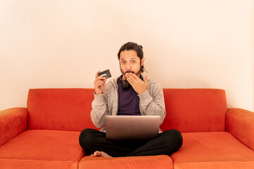 Fototapeta na wymiar Young man shopping online with credit card at home using computer sitting on an orange couch sofa