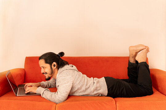 Young man dating online using the computer laptop laying on an orange couch sofa at home in the living room.