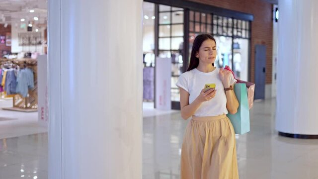 Young focused brunette woman in casual wear with paper bags in hand looking at map on modern smartphone while trying to find clothing store, using online navigation application during shopping in mall