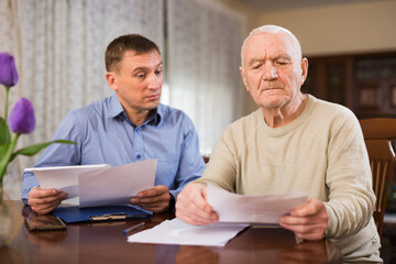 Adult man with his elderly father looking worriedly at papers analyzing their finances at home