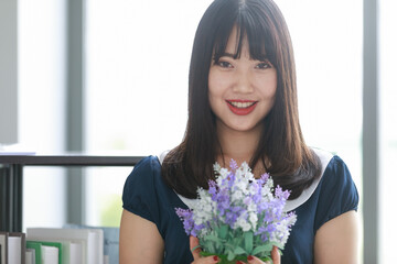 Vertical portrait shot of attractive smiling young Asian woman with long straight hairstyle in casual blue clothes holding the purple and white flowers while looking at the camera in the studio