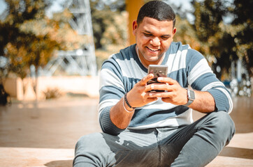 latin dominican man texting with his phone while smiling, sitting in the park - obrazy, fototapety, plakaty