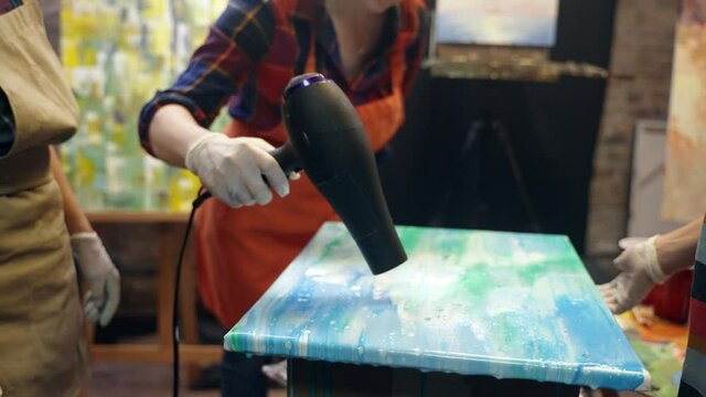 Women artists are drying acrylic paint on the canvas with fan. Artistic collaboration in workshop.