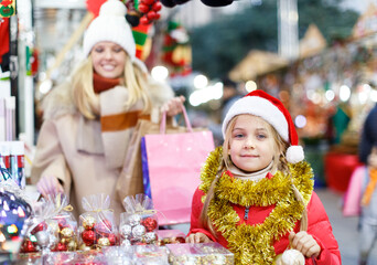 Portrait of smiling girl in Santa hat shopping on Christmas street fair with her mother on background