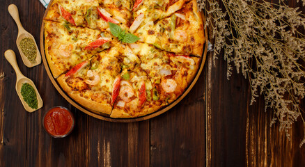 Vertical photo of homemade seafood pizza with shrimp, pineapples, bell pepper, crab sticks on wooden tray and table decorated with tomato sauce or ketchup, dried plants and oregano 