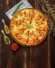 Vertical photo of homemade seafood pizza with shrimp, pineapples, bell pepper, crab sticks on wooden tray and table decorated with tomato sauce or ketchup, dried plants and oregano 