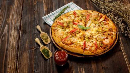 Selective focus and close up on homemade seafood pizza with shrimp, pineapples, bell pepper, crab sticks on wooden tray and table decorated with tomato sauce or ketchup, dried plants and oregano.
