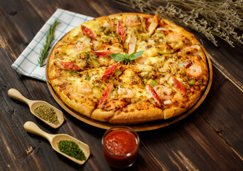 Selective focus and close up on homemade seafood pizza with shrimp, pineapples, bell pepper, crab sticks on wooden tray and table decorated with tomato sauce or ketchup, dried plants and oregano.