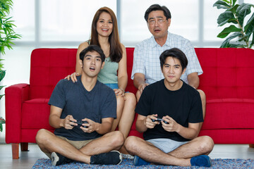 Four members Asian family consisting of parents, two adult sons spending time activity together, using joystick to enjoy videogames on sofa during holidays in cozy living room at home with happiness.