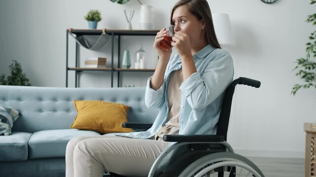 Slow motion of lonely young woman in wheelchair holding tea cup and drinking with serious face alone in apartment. People and special needs concept.