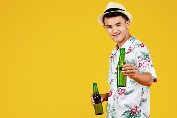 Young attractive Asian man in white Hawaiian shirt wearing white hat holding two bottles of beer and cheers against yellow background. Concept for beach vacation holiday