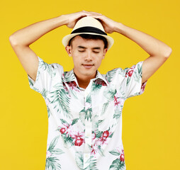 Young attractive Asian man in white Hawaiian shirt wearing white hat relaxing against yellow background. Concept for beach vacation holiday