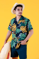 Young Asian man in colorful printed Hawaiian shirt smiles with happiness while holding swim ring. Half body studio portrait on yellow background. Summer holiday travel concept