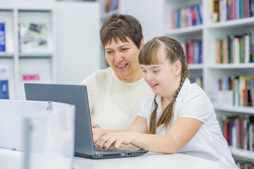 Happy senior woman teaches a young girl with down syndrome to use a laptop. Education for disabled children concept