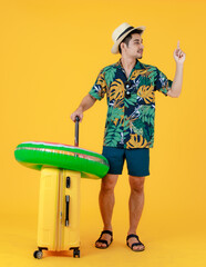 Profile shot, young Asian man in colorful Hawaiian shirt gives index finger up and pull yellow suitcase. Full body studio portrait on yellow background. Happy Summer holiday travel concept.