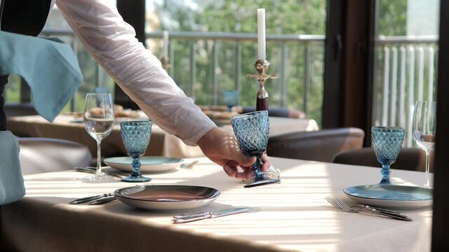 Waiter serves a table in an elite restaurant close-up. Men's hands arrange beautiful clean and empty glass glasses and plates.Decorative tableware and a candle holder.Restaurant with panoramic windows
