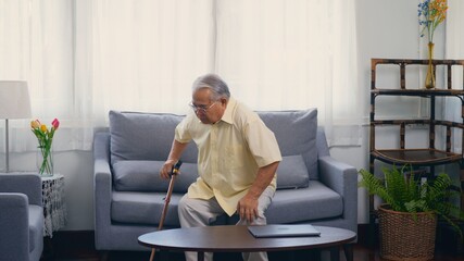 Old man grandfather with eyeglasses used a walking stick to lean the ground slowly stand up from the sofa carefully in living room at home. Asian senior old gray-haired suffering from knee pain ache