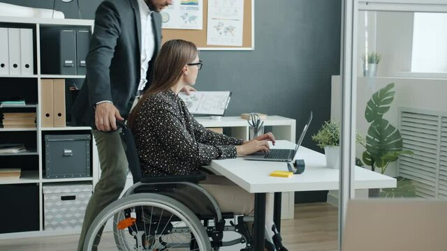 Slow motion of young businesswoman in wheelchair talking to coworker in office discussing business concentrated on job. Disability and employment concept.