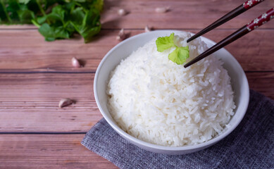 Steam organic white rice in a white ceramic bowl with chopsticks holding hot jasmine rice and apron and green coriander stained on a brown wooden table.