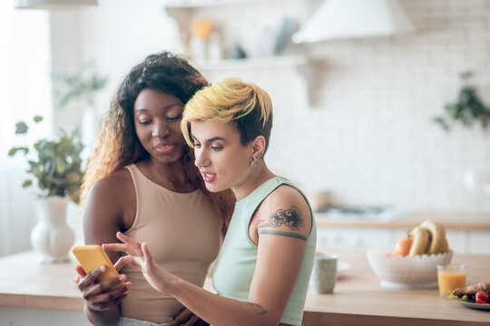 Dark-skinned woman holding smartphone caucasian girlfriend touching screen