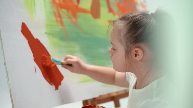 Girl with down syndrome draws with a brush on a large canvas in a white room, kid girl with special needs draws a color abstraction, 4k slow motion.