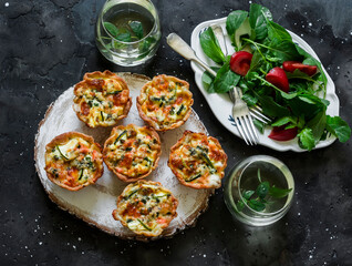 Aperitif table - mini quiche with salmon and zucchini, fresh tomato arugula salad and mint cocktail on a dark background, top view