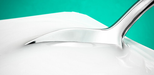 Yogurt cup and silver spoon on green background, white plastic container with yoghurt cream, fresh dairy product for healthy diet and nutrition balance.