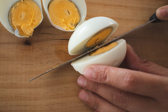 Person cutting a hard-boiled egg in half on a wooden surface next to another hard-boiled egg already cut with the yellow yolk in sight.