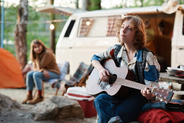 Young woman sitting outdoors and playing guitar during camping in the forest