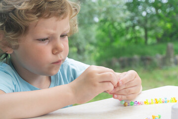 A little Caucasian girl, curly blonde, is captivated by creativity in the fresh air. The girl makes a homemade bead bracelet.