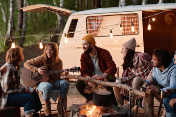 Group of friends sitting near the fire and playing guitar together during picnic on the nature