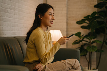 Side view of young happy Asian woman holding tv remote control changing channel while sitting on couch at home during late night