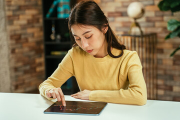 Successful young Asian female broker working and reading stock market graphs and data using digital tablet at home with concentration