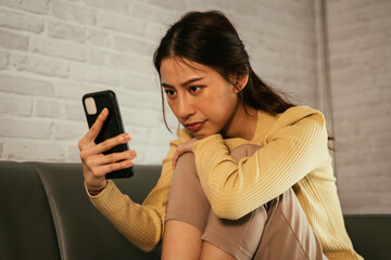 Young and tired Asian woman sitting anxiously and nervously on couch at home looking at mobile...