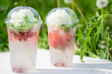 Cocktail with watermelon slices and ice cubes in a plastic cup, with a white flower. On a white wooden background, in the grass. Summer tropical refreshing drink.