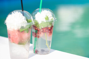 Cocktail with watermelon slices and ice in plastic cups with a flower. on a white background, on the edge of the resort pool. The concept of a luxury holiday. Outdoor pool background. Horizontal.