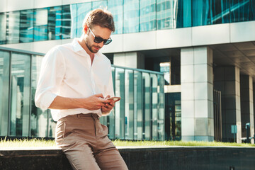 Handsome smiling stylish hipster lambersexual model.Modern man dressed in white shirt. Fashion male posing in the street near skyscrapers in sunglasses. Outdoors at sunset. Using smartphone apps