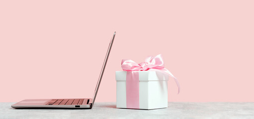 Laptop with white gift box with shining pink ribbon bow on pink background. Gift or holiday shopping concept. Mothers Day, birthday wedding or St Valentines day banner with copy space