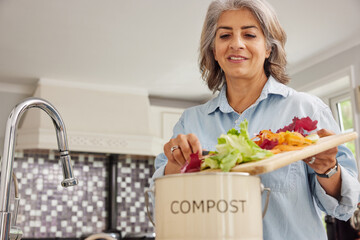 Mature Woman In Kitchen Making Compost Scraping Vegetable Leftovers Into Bin