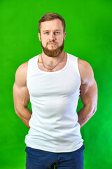 Fototapeta premium The red-haired man looks at the footage. Serious sportsman face with muscles. A Caucasian man with a beard stands on a green background in a white T-shirt and jeans.