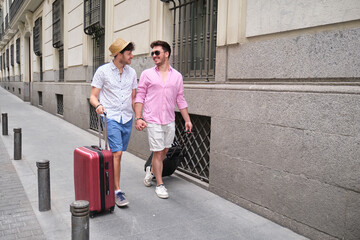 Gay couple carrying suitcases and going to the hotel holding hands.