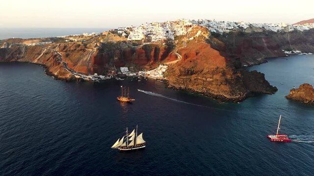 Aerial drone view video of beautiful sail boats cruising in the deep dark blue Aegean sea during sunset, Santorini, Cyclades, Greec