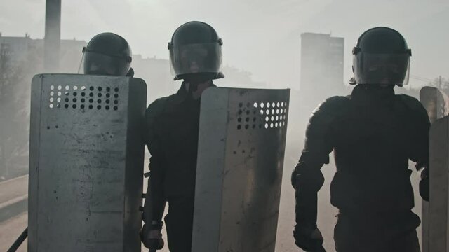 Slowmo tracking shot of three riot police officers in full gear carrying shields and batons and walking outdoors surrounded by smoke