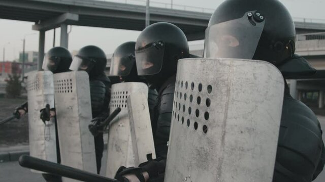 Tilt up slowmo shot of unrecognizable riot police officers in full gear blocking street and beating shields with batons