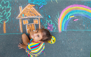 The child draws a house and a rainbow on the asphalt with chalk. Selective focus.